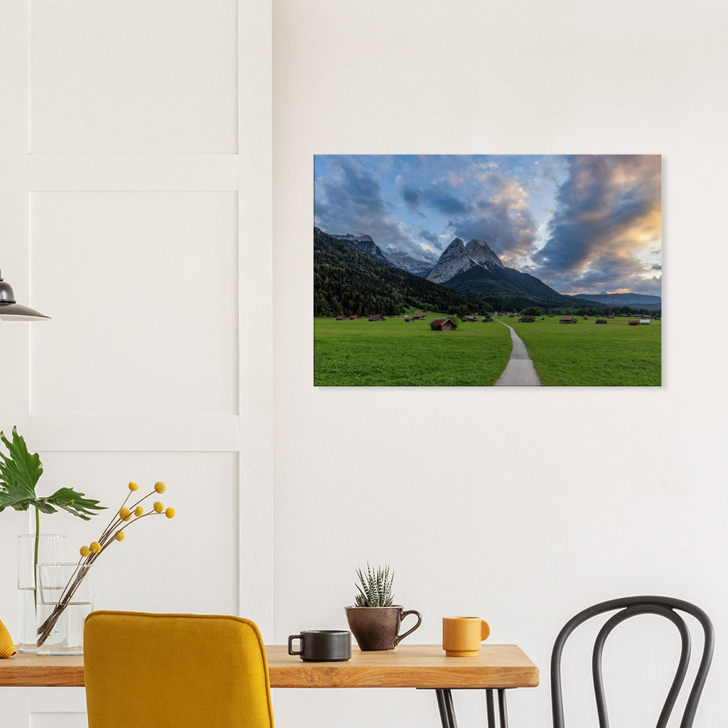 Landschaftsfotografie aus Garmisch-Partenkirchen mit Blick auf das imposante Wettersteingebirge. Ein schmaler Weg führt durch weitläufige grüne Wiesen mit traditionellen Heustadeln. Dramatische Wolken und sanftes Abendlicht tauchen die bayerische Alpenlandschaft in eine stimmungsvolle Atmosphäre.