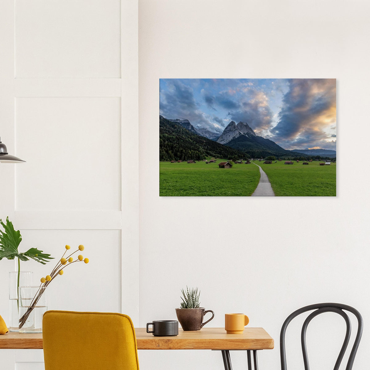 Landschaftsfotografie aus Garmisch-Partenkirchen mit Blick auf das imposante Wettersteingebirge. Ein schmaler Weg führt durch weitläufige grüne Wiesen mit traditionellen Heustadeln. Dramatische Wolken und sanftes Abendlicht tauchen die bayerische Alpenlandschaft in eine stimmungsvolle Atmosphäre.