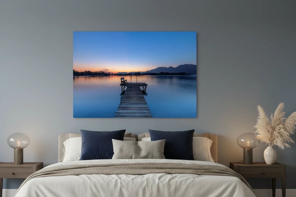 Aluminium-Print mit Sonnenaufgang am Kochelsee: Ruhiger See in sanften Blau- und Orangetönen, ein hölzerner Steg mit Bank führt ins stille Wasser, im Hintergrund Berge und die zarte Morgenstimmung des beginnenden Tages.