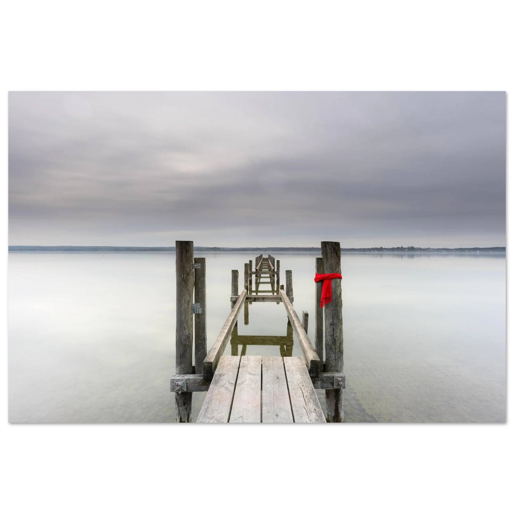 Alter Holzsteg am Ammersee mit einem leuchtend roten Schal, der an einem Pfosten befestigt ist. Die ruhige Langzeitbelichtung, das glatte Wasser und der sanft bewölkte Himmel schaffen eine stille, minimalistische Atmosphäre in Oberbayern.
