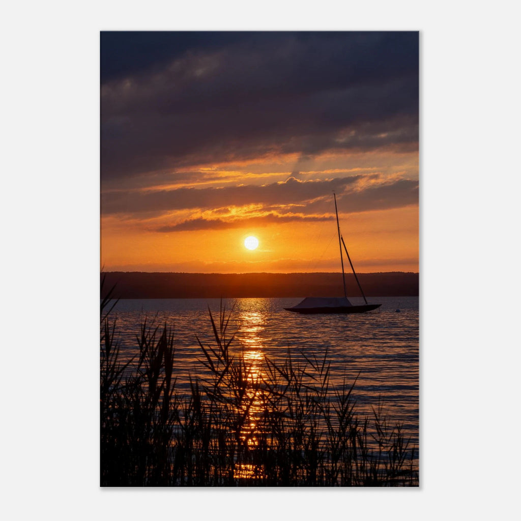 Vertikales Leinwandbild mit einem leuchtenden Sommer-Sonnenuntergang am Ammersee in Bayern. Goldene und orangefarbene Lichtstimmungen spiegeln sich auf dem Wasser, während ein Segelboot ruhig auf dem See liegt und feines Schilf im Vordergrund die natürliche Atmosphäre abrundet.