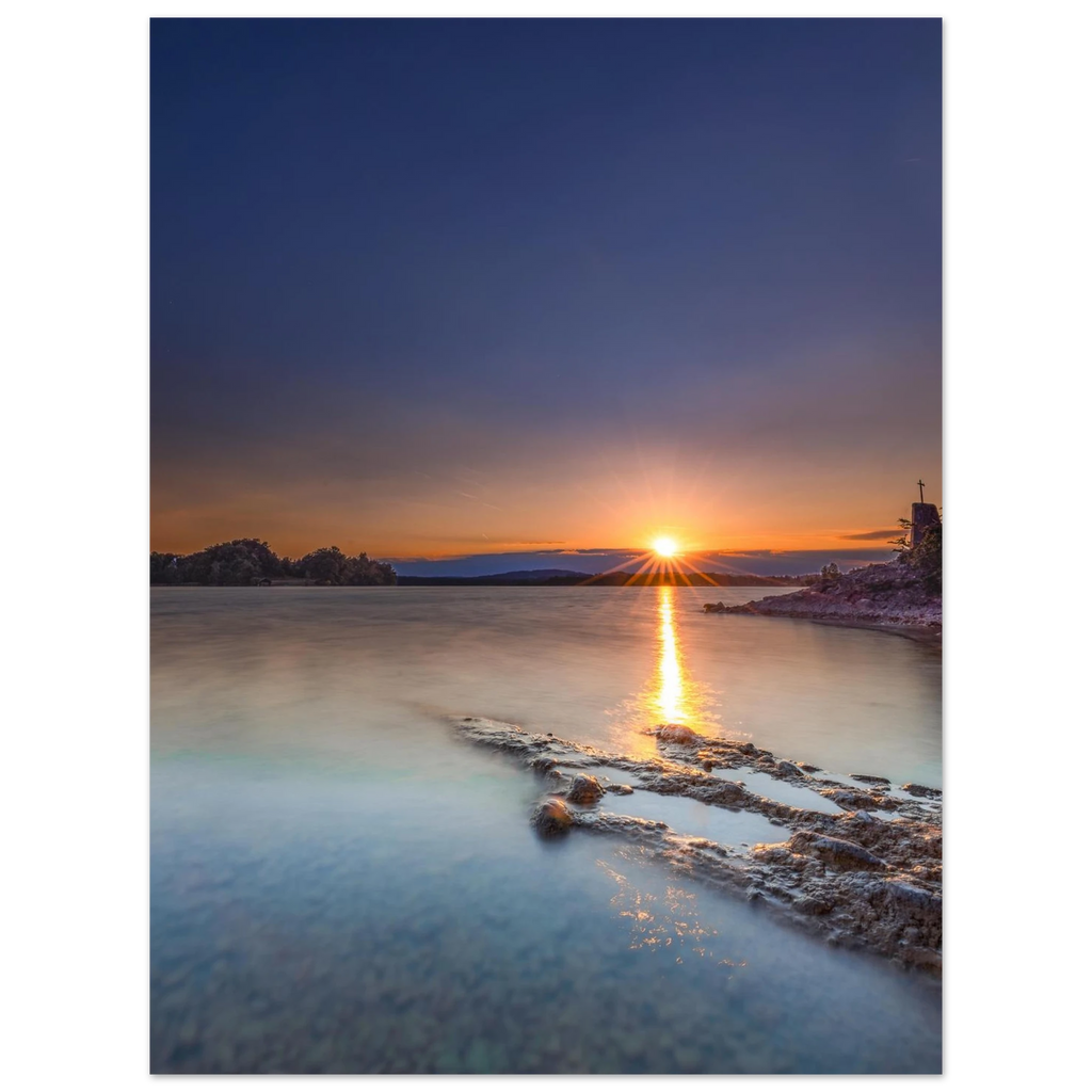 Sommerlicher Sonnenuntergang am Staffelsee in Oberbayern. Die tief stehende Sonne berührt den Horizont und spiegelt sich als goldener Lichtstreifen auf dem ruhigen, durch Langzeitbelichtung weich wirkenden Wasser. Im Vordergrund ragen strukturierte Felsen ins klare Uferwasser, rechts ist eine kleine Kapelle auf einer Landzunge zu sehen. Der Himmel leuchtet in warmen Orange- und kühlen Blautönen und vermittelt eine friedliche Abendstimmung.