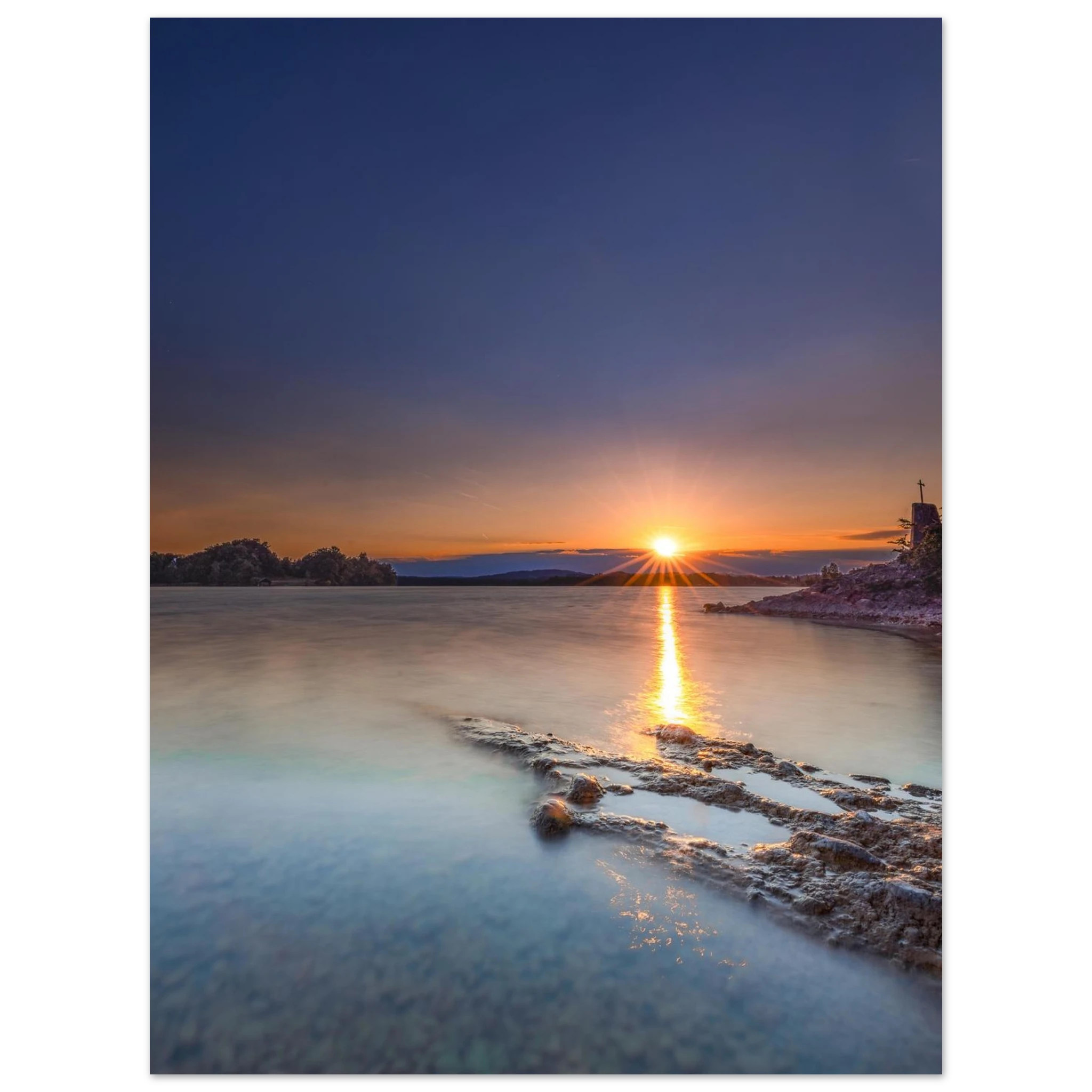 Sommerlicher Sonnenuntergang am Staffelsee in Oberbayern. Die tief stehende Sonne berührt den Horizont und spiegelt sich als goldener Lichtstreifen auf dem ruhigen, durch Langzeitbelichtung weich wirkenden Wasser. Im Vordergrund ragen strukturierte Felsen ins klare Uferwasser, rechts ist eine kleine Kapelle auf einer Landzunge zu sehen. Der Himmel leuchtet in warmen Orange- und kühlen Blautönen und vermittelt eine friedliche Abendstimmung.
