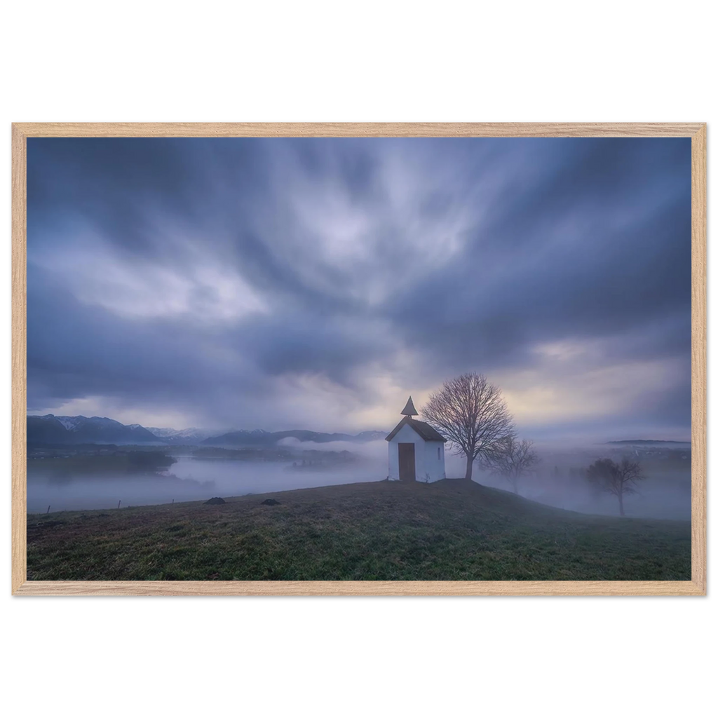 Hoch über der Landschaft bei Aidling im Blauen Land steht die kleine Mesnerhauskapelle – still, schlicht und eingebettet in einen Morgen, der kaum leiser sein könnte. Nebelschleier ziehen durch die Landschaft, während sich das erste Licht vorsichtig seinen Weg durch Wolken und Himmel bahnt. Die Weite öffnet den Blick, die Zeit scheint für einen Augenblick langsamer zu werden.
Poster mit Holzrahmen