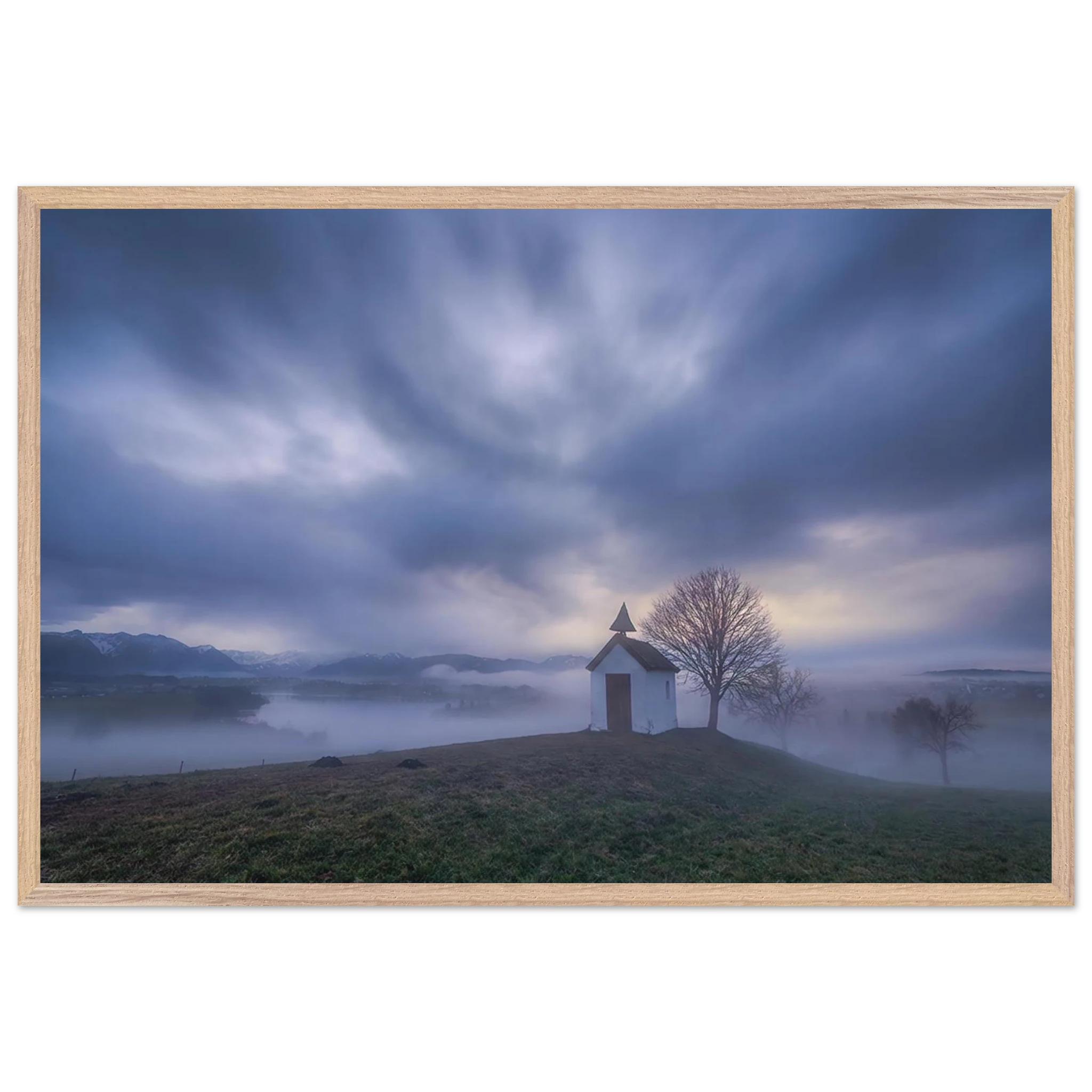 Hoch über der Landschaft bei Aidling im Blauen Land steht die kleine Mesnerhauskapelle – still, schlicht und eingebettet in einen Morgen, der kaum leiser sein könnte. Nebelschleier ziehen durch die Landschaft, während sich das erste Licht vorsichtig seinen Weg durch Wolken und Himmel bahnt. Die Weite öffnet den Blick, die Zeit scheint für einen Augenblick langsamer zu werden.
Poster mit Holzrahmen