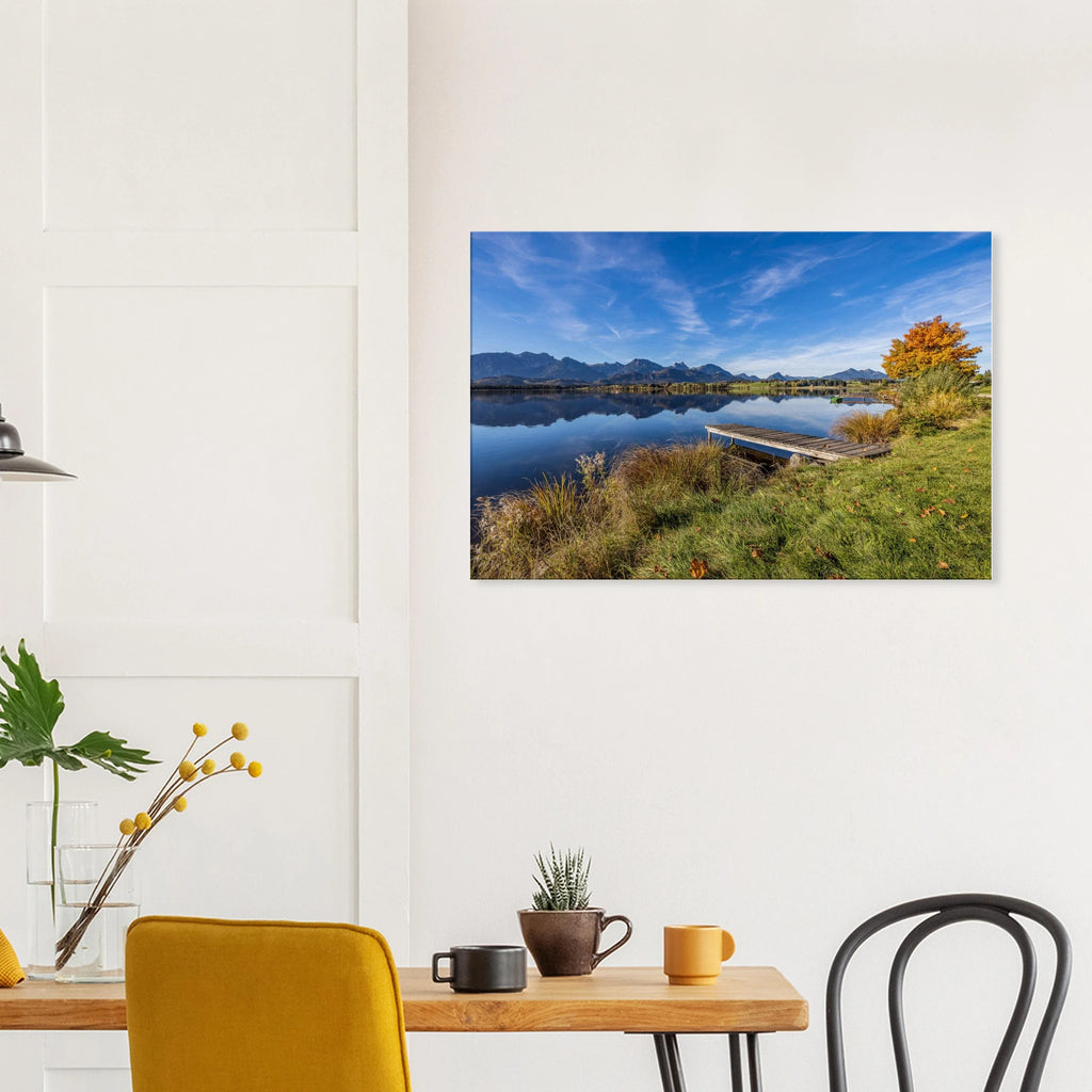 Ein goldener Herbsttag im Allgäu. Der Hopfensee liegt still vor der beeindruckenden Kulisse der Berge, das klare Wasser trägt ihre Spiegelungen und am Ufer leuchtet der Herbst in warmen Farben. Der kleine Holzsteg führt hinaus in diese besondere Ruhe, die man manchmal nur in der Natur findet.