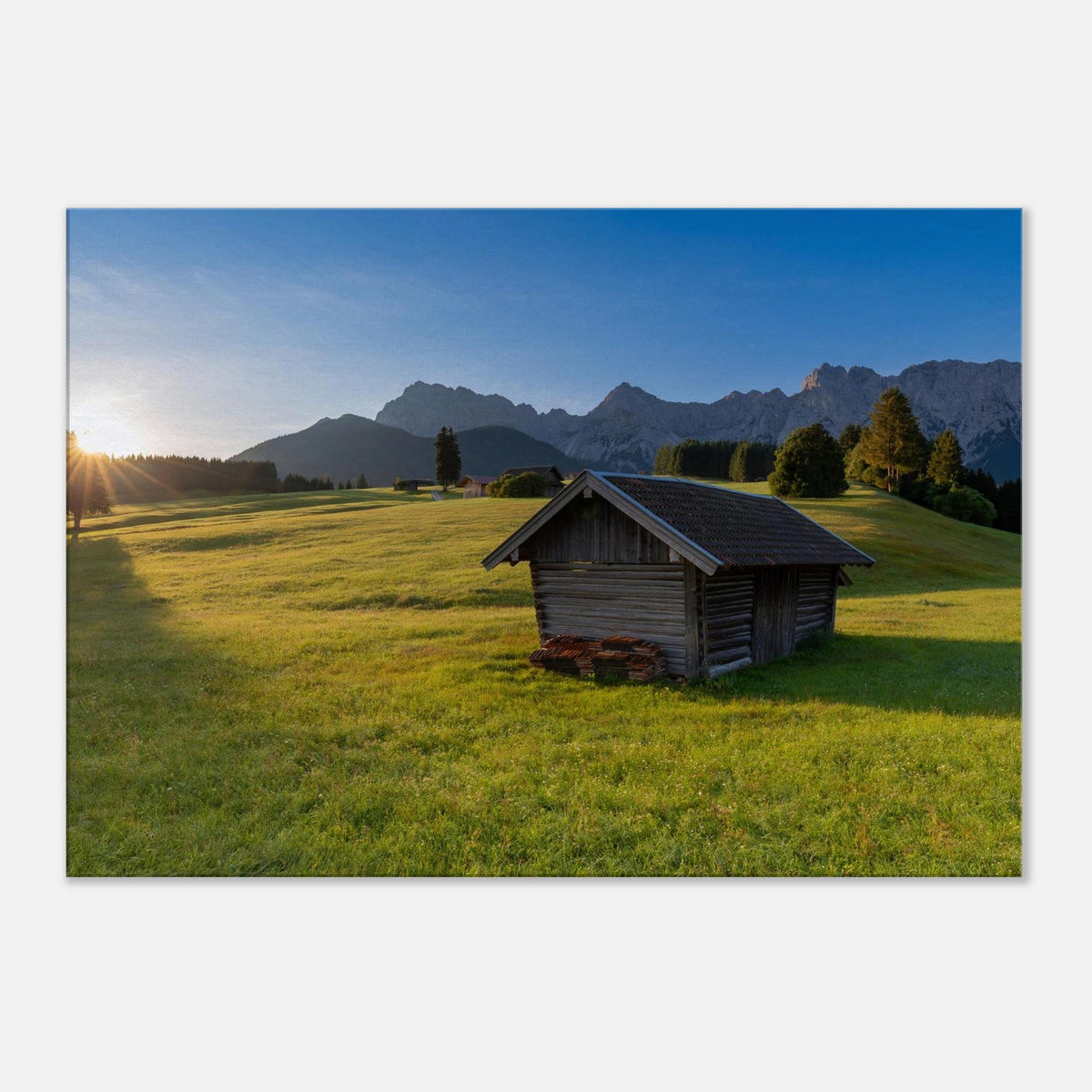 Leinwandbild mit traditioneller Holzhütte auf einer sonnigen, grünen Almwiese vor der beeindruckenden Bergkulisse des Karwendelgebirges. Warmes Morgenlicht taucht die idyllische Alpenlandschaft in eine natürliche, ruhige Stimmung.
