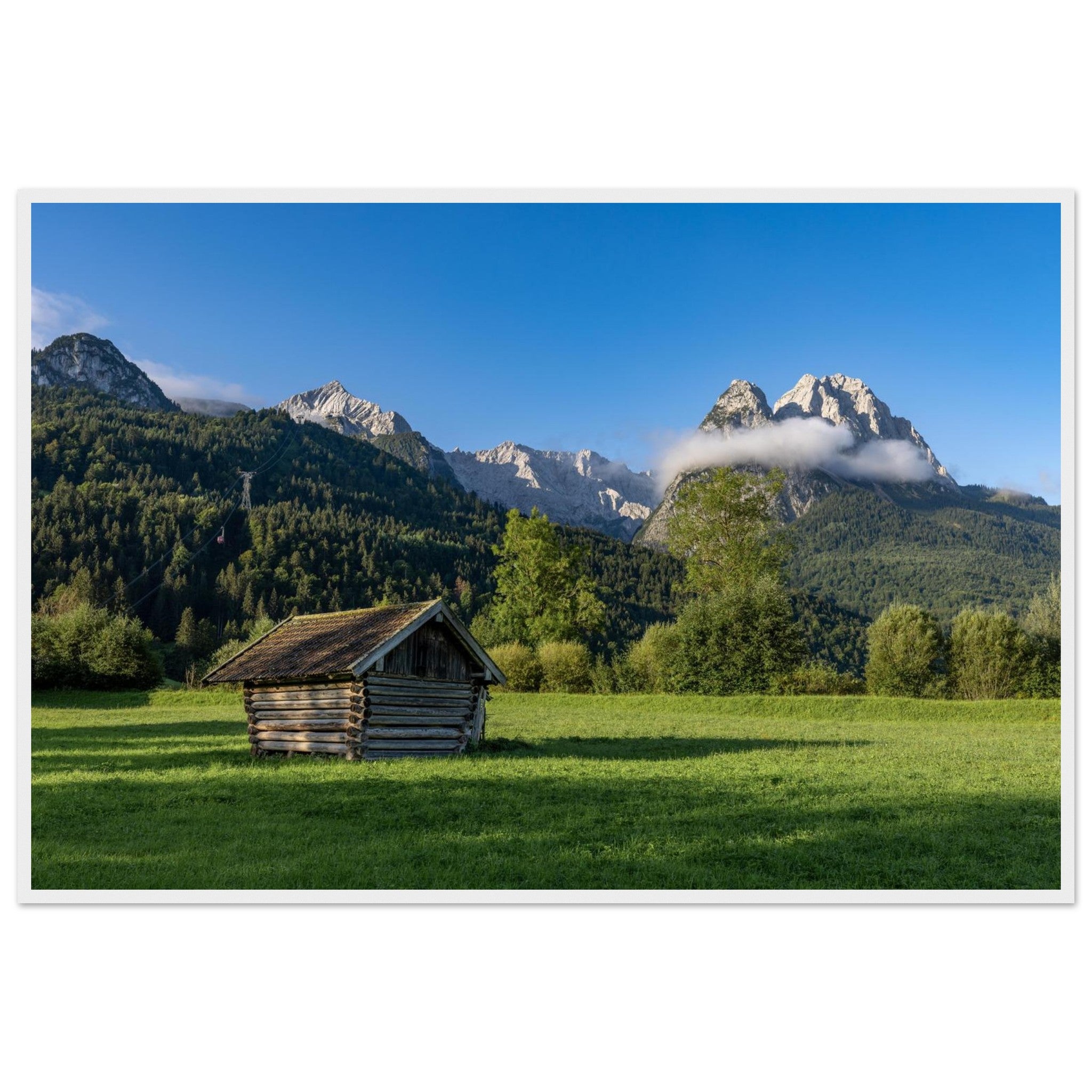 Gerahmtes Landschaftsposter mit weissem Holzrahmen im Format 60x90 cm  Das Motiv zeigt eine kleine Holzhütte auf einer Wiese vor beeindruckender Bergkulisse unter blauem Himmel – eingebettet in natürliche Materialien und eine ruhige, harmonische Wohnatmosphäre.
