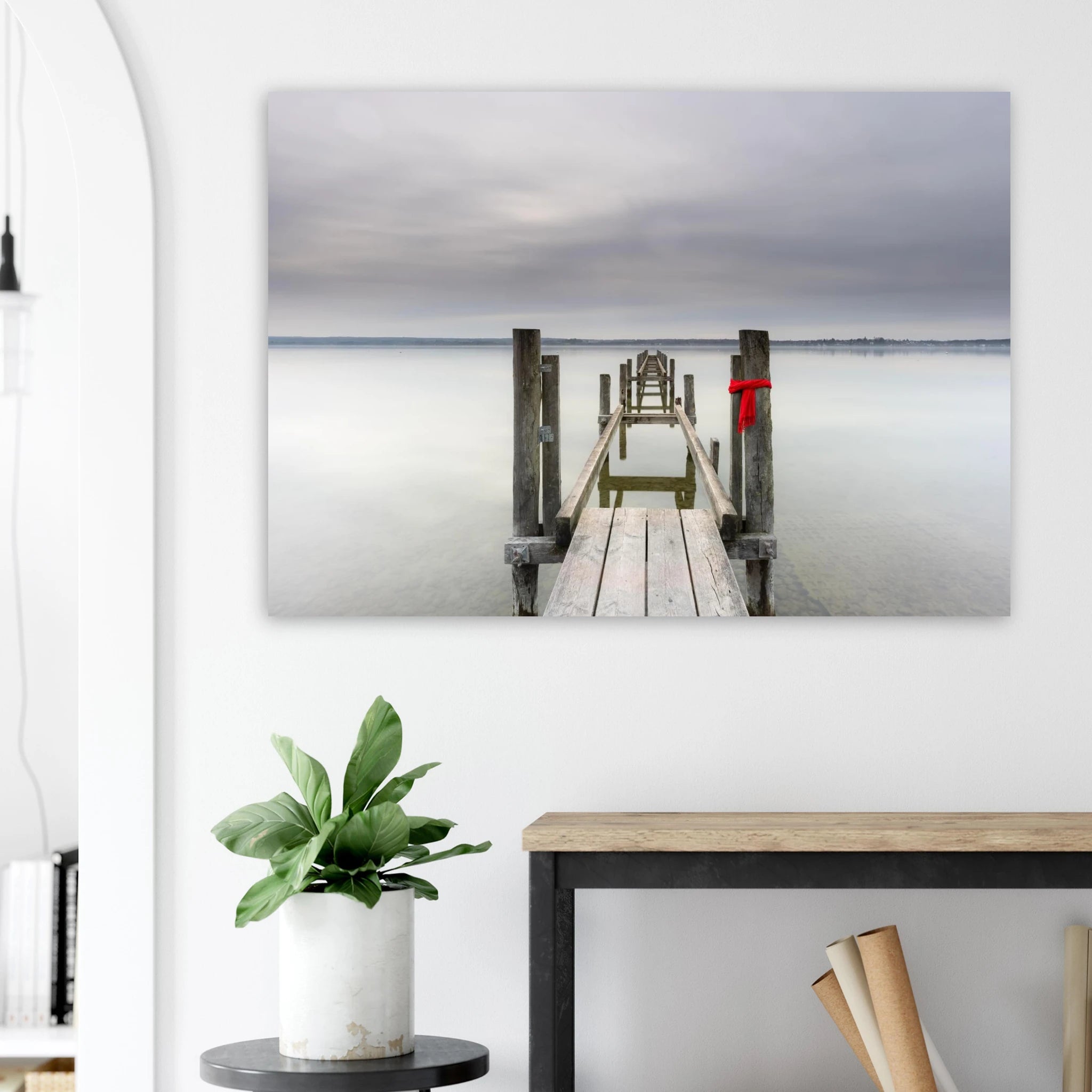 Alter Holzsteg am Ammersee mit einem leuchtend roten Schal, der an einem Pfosten befestigt ist. Die ruhige Langzeitbelichtung, das glatte Wasser und der sanft bewölkte Himmel schaffen eine stille, minimalistische Atmosphäre in Oberbayern.