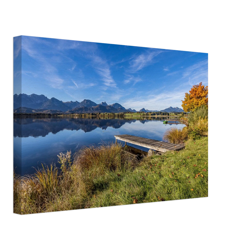 Ein goldener Herbsttag im Allgäu. Der Hopfensee liegt still vor der beeindruckenden Kulisse der Berge, das klare Wasser trägt ihre Spiegelungen und am Ufer leuchtet der Herbst in warmen Farben. Der kleine Holzsteg führt hinaus in diese besondere Ruhe, die man manchmal nur in der Natur findet. Foto auf Leinwand 